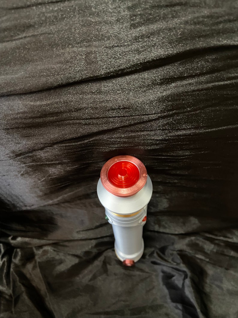 K&ouml;nnte beinhalten: Ein wei&szlig;es Lichtschwert mit einem roten, kupfernen und goldenen Griff. Das Lichtschwert liegt auf einem schwarzen Stoffhintergrund.