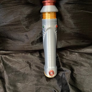 K&ouml;nnte beinhalten: Ein silberner Lichtschwertgriff mit goldenen und roten Akzenten. Der Griff hat eine strukturierte Oberfl&auml;che und einen runden, roten Knopf unten.