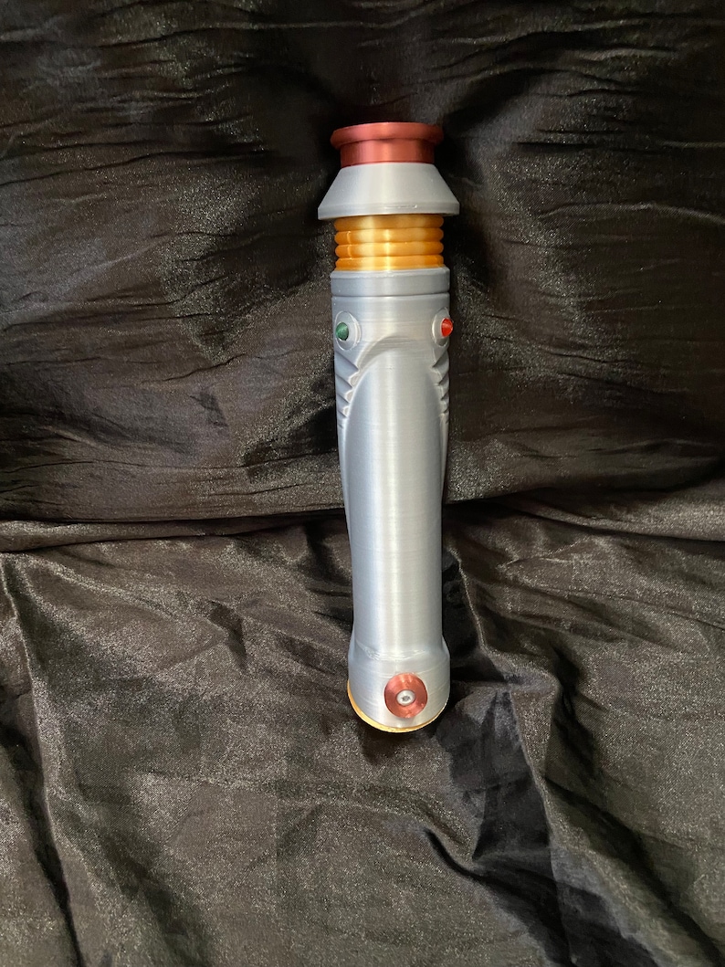 K&ouml;nnte beinhalten: Ein silberner Lichtschwertgriff mit goldenen und roten Akzenten. Der Griff hat eine strukturierte Oberfl&auml;che und einen runden, roten Knopf unten.