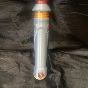 K&ouml;nnte beinhalten: Ein silberner Lichtschwertgriff mit goldenen Akzenten und roten und gr&uuml;nen Kn&ouml;pfen. Der Griff hat ein kupferfarbenes Ober- und Unterteil.