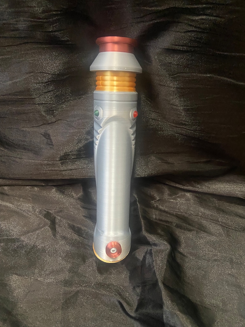 K&ouml;nnte beinhalten: Ein silberner Lichtschwertgriff mit goldenen Akzenten und roten und gr&uuml;nen Kn&ouml;pfen. Der Griff hat ein kupferfarbenes Ober- und Unterteil.