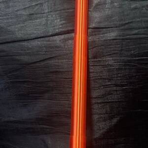 K&ouml;nnte beinhalten: Ein rotes und oranges Lichtschwert-Spielzeug mit einem silbernen Griff und goldenen Akzenten. Das Lichtschwert befindet sich auf einer schwarzen Oberfl&auml;che.