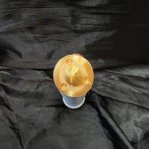 K&ouml;nnte beinhalten: Ein goldfarbener Metalldeckel mit einer kleinen, runden, erhabenen Mitte und drei kleinen, runden Vertiefungen am Rand.