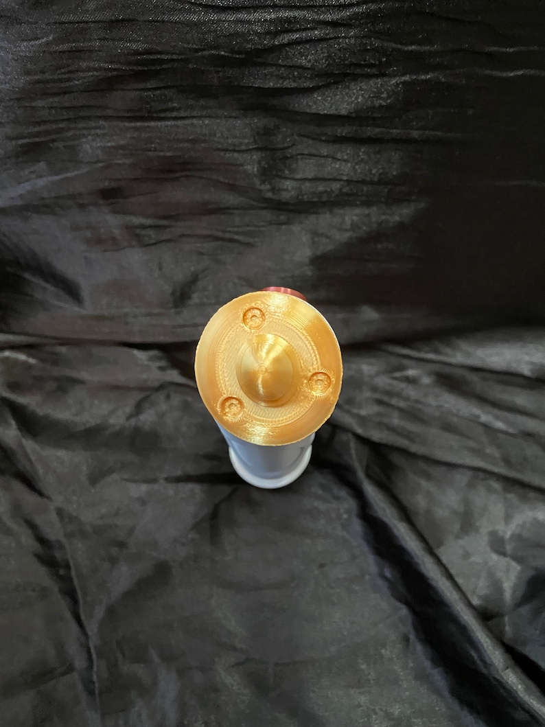 K&ouml;nnte beinhalten: Ein goldfarbener Metalldeckel mit einer kleinen, runden, erhabenen Mitte und drei kleinen, runden Vertiefungen am Rand.