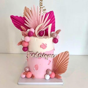 Peut inclure: Gâteau rose à deux étages décoré de éventails en papier rose et pêche, des chiffres en bois "61" et le nom "Frederique" en lettres en bois. Le gâteau est surmonté de boules de bonbons roses et blanches.