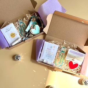 Peut inclure: Deux boîtes cadeaux en carton ouvertes remplies de copeaux de bois, contenant chacune un marque-page en acrylique transparent avec une icône de livre, un charm en forme de cœur et une carte. Une carte indique Merci pour cette année, l'autre merci maitresse de m'avoir aidé à grandir.