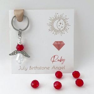 May include: A silver angel charm with a red crystal birthstone. The charm is attached to a silver key ring. The card reads "Ruby July Birthstone Angel".