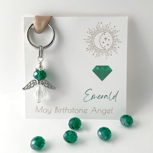 Peut inclure: Un charme d'ange en argent avec des ailes en cristal vert et une tête en cristal vert. Le charme est attaché à un porte-clés en argent. Une carte avec le texte "Emerald May Birthstone Angel" est en arrière-plan. Il y a plusieurs perles en cristal vert éparpillées autour du charme.