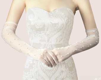 May include: White bridal gloves with pearl embellishments. The gloves are made of sheer fabric and extend to the elbow. The gloves are worn by a person with a white dress.