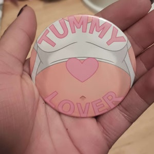May include: A pink and white button with the text "TUMMY LOVER" in pink letters. A pink heart is in the center of the button.
