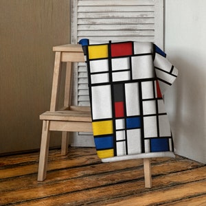 May include: A white fleece throw blanket with a geometric pattern of black lines and squares filled with red, yellow, and blue. The blanket is draped over a wooden step stool.