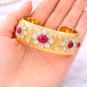May include: A gold cuff bracelet featuring a brushed texture finish, adorned with three prominent oval-shaped red gemstones. Each red stone is surrounded by a cluster of small, clear, round-cut white stones, with additional scattered white stones.