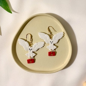 以下が含まれることがあります： 白いフクロウのイヤリングのペア。翼を広げ、それぞれ小さな赤い封筒を持っています。イヤリングは、薄い黄色の楕円形のお皿に飾られています。フクロウの目は黒色です。