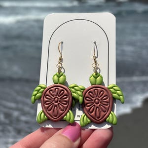 Puede incluir: Pendientes de tortuga hechos a mano. Cada pendiente presenta un caparazón marrón con un diseño floral y apéndices verdes en forma de aleta. Los pendientes cuelgan de ganchos dorados. El fondo muestra una playa y el océano.