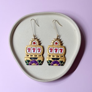 May include: A pair of gold-colored slot machine earrings with the number 777 displayed. The earrings have red, green, purple, and white accents. The earrings are displayed on a light gray, oval-shaped plate against a light purple background.