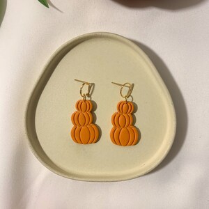 Puede incluir: Un par de pendientes colgantes de color naranja en forma de calabaza con aros dorados. Los pendientes están apilados uno encima del otro para crear un efecto tridimensional.
