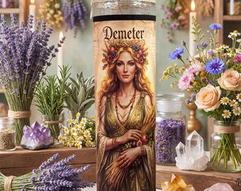 Może przedstawiać: Wysoka cylindryczna szklana świeca z nadrukowaną ilustracją bogini Demeter z długimi blond włosami, wiankiem z kwiatów i snopem pszenicy. Etykieta zawiera napis Demeter wykonany ozdobną czcionką w górnej części produktu.