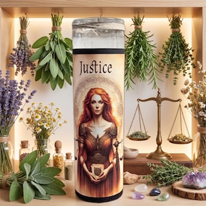 May include: A cylindrical glass candle featuring a label with the word Justice and an illustration of a red-haired figure in armor holding a scale. The candle is surrounded by bundles of dried herbs, a brass balance scale, and small crystals.