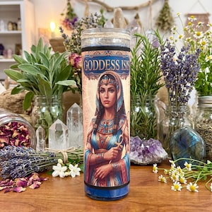 A 7 day glass candle featuring an illustration of Goddess Isis, surrounded by jars of herbs, lavender, crystals, and flowers on a wooden surface.