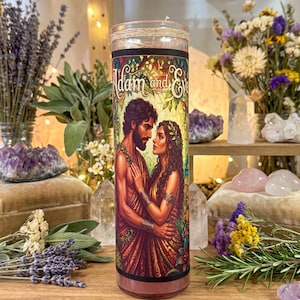 May include: A cylindrical glass candle featuring a label with the text Adam and Eve, depicting a man and a woman embracing in a lush garden setting. The candle is topped with a thin twine wrap and contains a solid wax core within the clear glass vessel.
