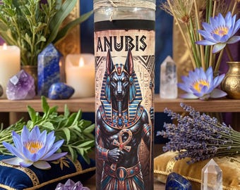 May include: A tall cylindrical glass candle featuring a graphic print of the Egyptian deity Anubis with the text ANUBIS at the top. The figure is depicted in blue and gold attire holding an ankh, set against a background of hieroglyphics.