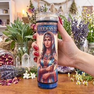 A hand with long red fingernails holding a 7 day glass candle featuring an illustration of Goddess Isis, surrounded by jars of herbs, lavender, crystals, and flowers on a wooden surface.
