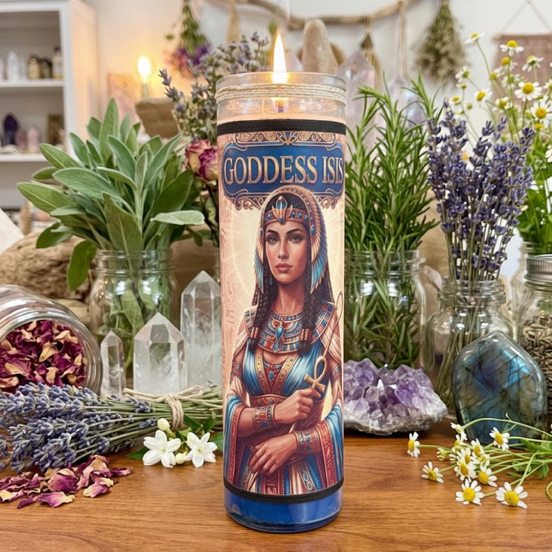 A lit 7 day glass candle featuring an illustration of Goddess Isis, surrounded by jars of herbs, lavender, crystals, and flowers on a wooden surface.