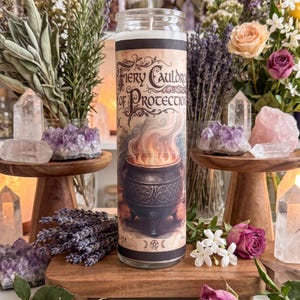 May include: A tall cylindrical glass candle featuring a label with the text Fiery Cauldron of Protection. The label displays an illustration of a black iron cauldron with Celtic knotwork patterns and orange flames rising from the top.