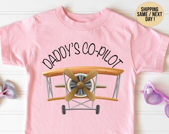 May include: Pink t-shirt with a graphic of a vintage airplane and the text "Daddy's Co-Pilot".