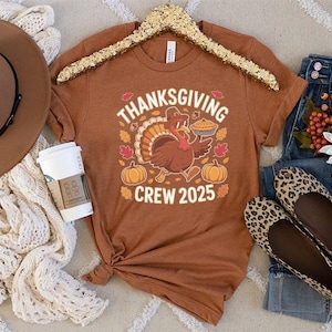 May include: A rust-colored t-shirt with a Thanksgiving graphic featuring a turkey carrying a pie, pumpkins, and the text "Thanksgiving Crew 2025." The image also includes a brown hat, a white scarf, jeans, and leopard print flats.