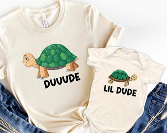 May include: Two cream-colored shirts with turtle graphics. One is a t-shirt with the word "DUUUDE" below the turtle. The other is a baby onesie with the words "LIL DUDE" below a similar turtle graphic. Blue jeans are visible.