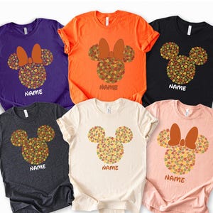 May include: A collection of six short-sleeved t-shirts in various colors: purple, orange, black, charcoal gray, cream, and pink. Each shirt features a Minnie Mouse head design made of autumn leaves, with a bow and the word "NAME" printed below.