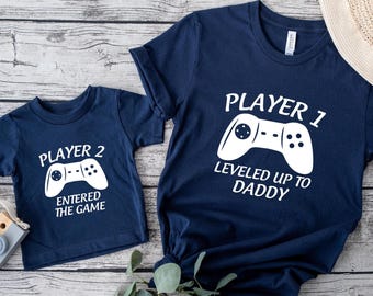 May include: Two navy blue t-shirts with white text and graphics. One shirt says "Player 1, Leveled Up to Daddy" with a controller graphic. The other says "Player 2, Entered the Game" with a controller graphic. The shirts are on a wooden surface.