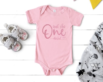 May include: Pink baby onesie with the text "isn't she one derful" in pink glitter lettering.