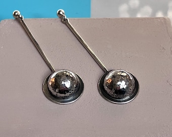 May include: A pair of silver earrings. Each earring features a long, thin post with a small ball at the top. The bottom of the post connects to a hammered, spherical design set within a circular frame. The earrings are displayed on a light purple surface.