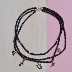May include: A black beaded necklace with four silver charms, including a heart, a foot, a cross, and a hand. The necklace is layered and has a silver clasp.