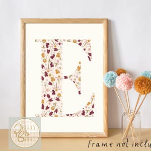 May include: A framed print of the letter "E" with a floral design in burgundy, yellow, and cream. The print is in a light wood frame. Decorative pom-poms in various colours are in a glass jar.