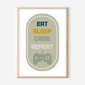 May include: A framed print with a green oval background and a white border. The oval contains the text "Eat Sleep Game Repeat" in a pixelated font with the words "Eat" and "Repeat" in dark blue, "Sleep" in gold, and "Game" in grey. A grey video game controller is centred below the text.