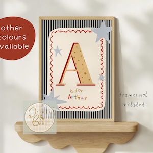 May include: A framed print with the letter "A" and the text "is for Arthur." The letter is red with gold star accents, set against a cream background with a red scalloped border. Blue star cutouts and a navy striped background complete the design.