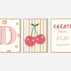 May include: Three printable wall art prints with a pink and yellow colour scheme. The first print features the letter D with a pink background and yellow flowers. The second print features two red cherries with happy faces on a striped background. The third print features the words "CREATE learn READ Explore" in pink, yellow, and green.