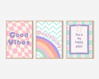 May include: Three pastel-colored prints with motivational quotes. The first print has a pink and white checkered background with the text "Good Vibes". The second print has a green and white chevron pattern with a rainbow and the text "Positivity". The third print has a green and white striped background with a purple frame and the text "This is my happy place".