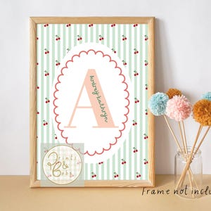 May include: A framed print with a light pink letter "A" and the name "Audrey Kathryn". The background has a green and white striped pattern with red cherry illustrations. A small logo is in the bottom left corner.