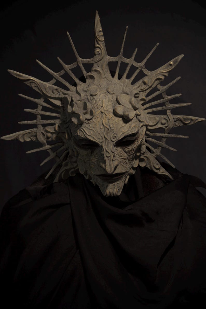 May include: A detailed, ornate mask with a crown-like structure. The mask is a light, neutral color with intricate carvings and a stern expression. The mask is set against a dark background, with a black fabric draped below.