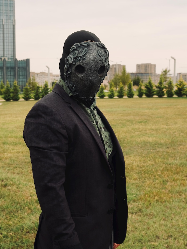 May include: A person wearing a black suit jacket and a black mask with intricate details. The mask has two eye holes and a floral design. The person is standing in a grassy field with buildings in the background.