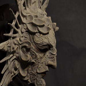 May include: A detailed, gray-toned mask with an intricate design, featuring a prominent nose and detailed facial features. The mask has a crown-like structure and is set against a dark background, suggesting a theatrical or fantasy theme.