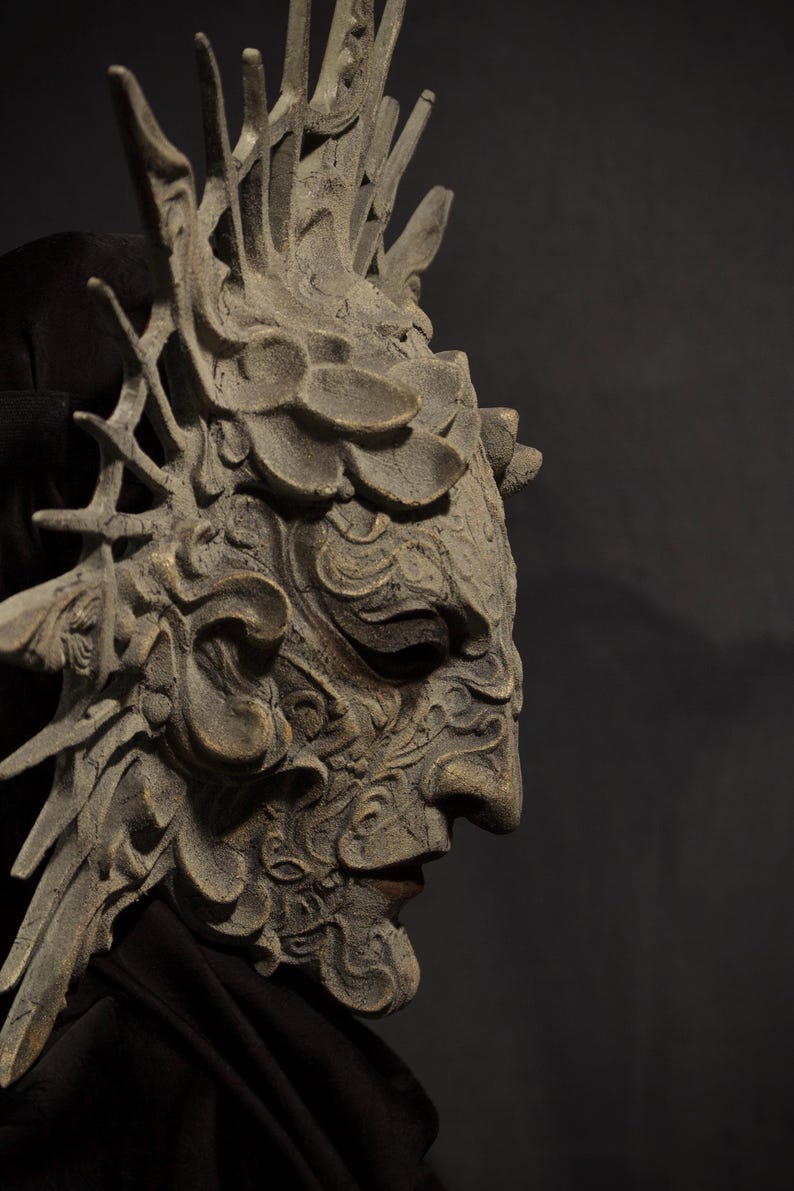 May include: A detailed, gray-toned mask with an intricate design, featuring a prominent nose and detailed facial features. The mask has a crown-like structure and is set against a dark background, suggesting a theatrical or fantasy theme.