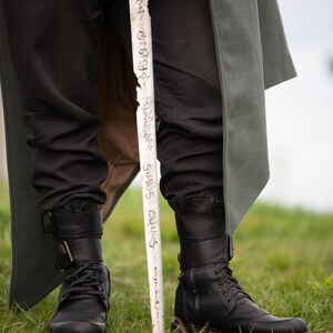 Op de afbeelding: Zwarte leren laarzen met gespen en veters, gecombineerd met een zwarte broek. Een witte staf met tekst wordt voor de benen gehouden. Een lange, grijze jas is zichtbaar op de achtergrond. De sc&egrave;ne speelt zich af op groen gras.