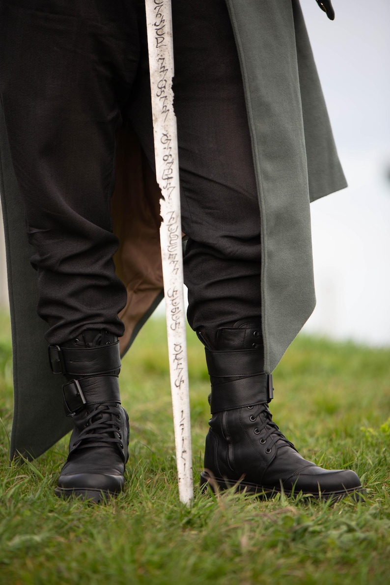 Op de afbeelding: Zwarte leren laarzen met gespen en veters, gecombineerd met een zwarte broek. Een witte staf met tekst wordt voor de benen gehouden. Een lange, grijze jas is zichtbaar op de achtergrond. De sc&egrave;ne speelt zich af op groen gras.