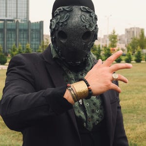 May include: A person wearing a black suit jacket and a black mask with intricate floral designs. The mask covers the person's face and has two eye holes. The person is wearing a gold bracelet and a ring on their finger.