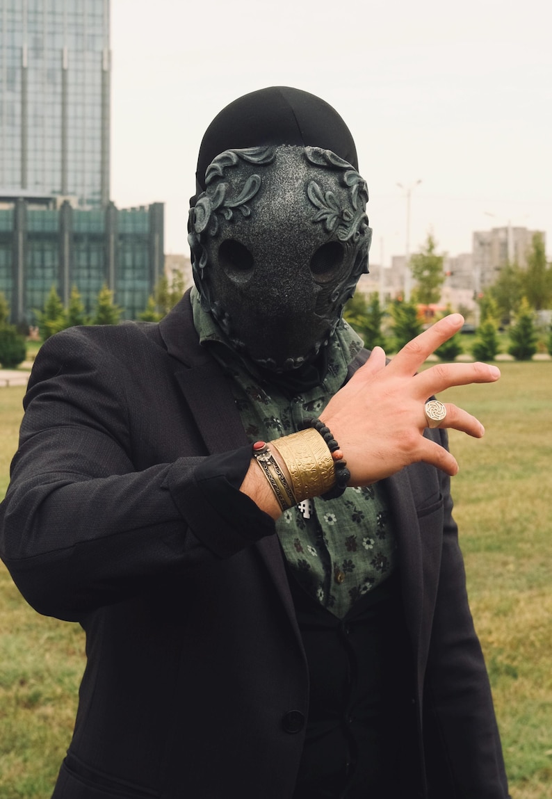 May include: A person wearing a black suit jacket and a black mask with intricate floral designs. The mask covers the person's face and has two eye holes. The person is wearing a gold bracelet and a ring on their finger.